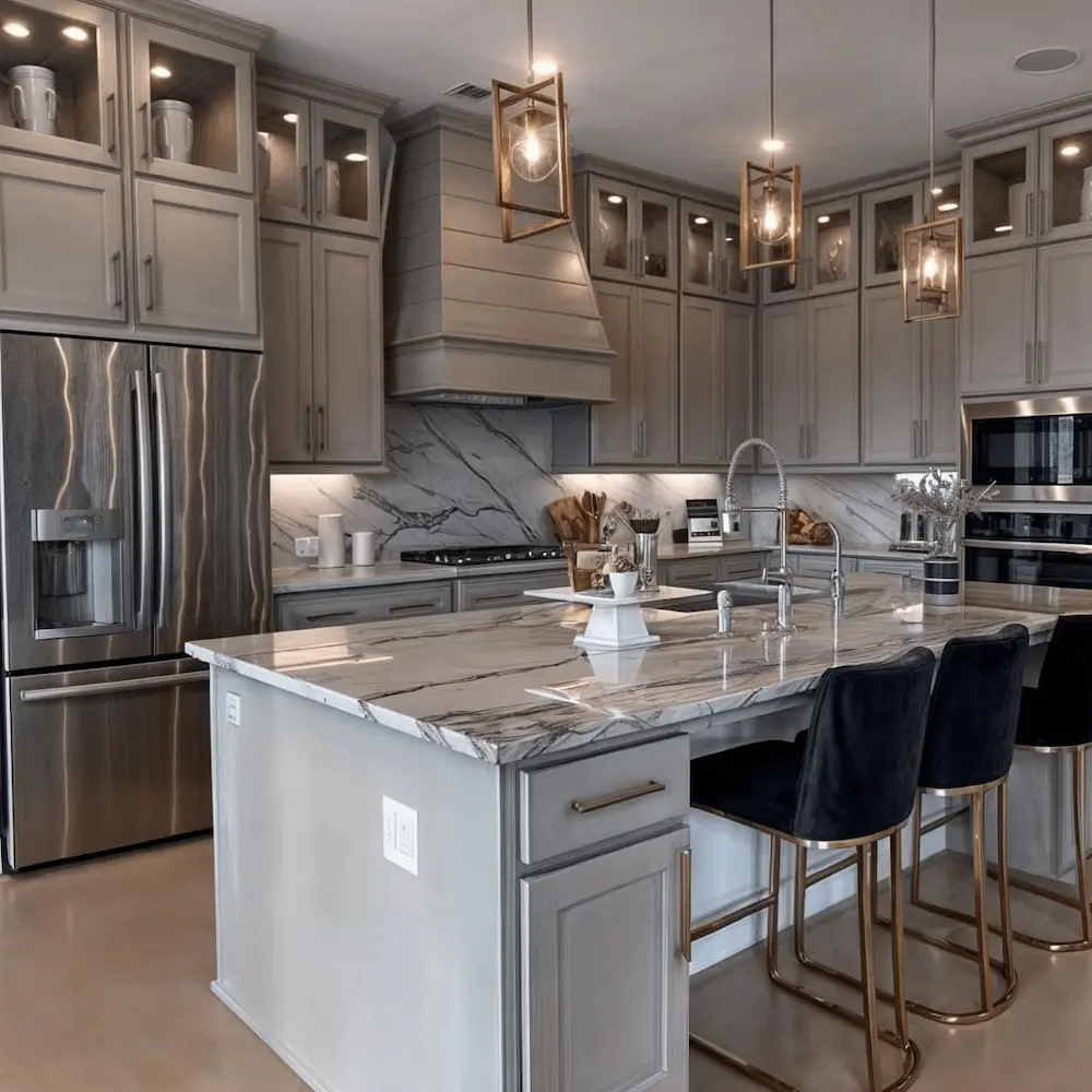 Transitional greige shaker kitchen with illuminated stacked millwork, continuous stone slab backsplash, bold-veined island, and open-frame brass pendants in Pelham, Westchester County — JMR Construction